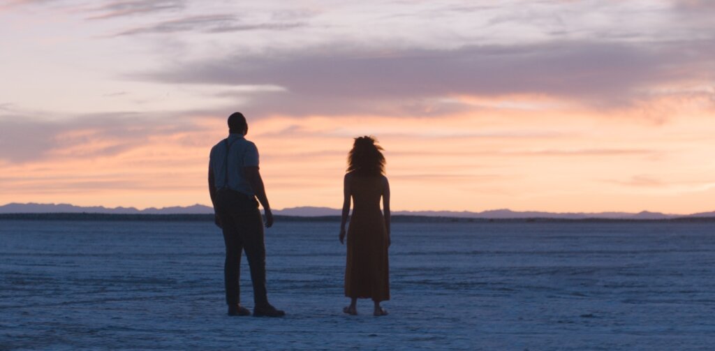 Will and Emma face each other on a salt flat at sunset in Nine Days