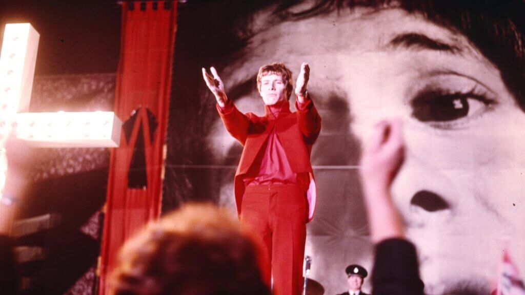 

Steven Shorter addresses a crowd in a red suit beneath an illuminated cross and a giant projected face in Privilege.