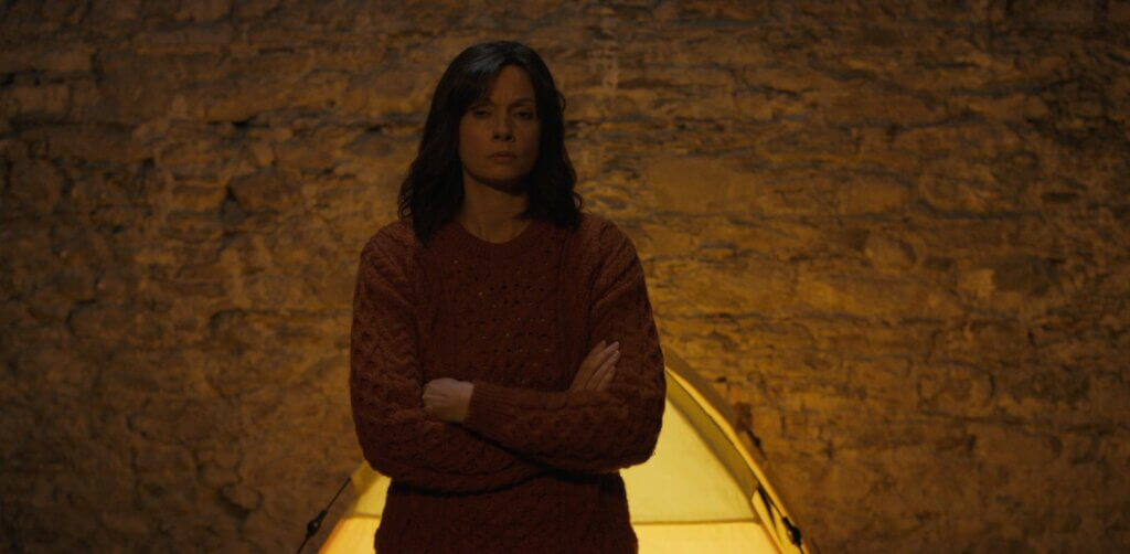 Dani stands alone against the illuminated stone wall of her remote house in Oddity