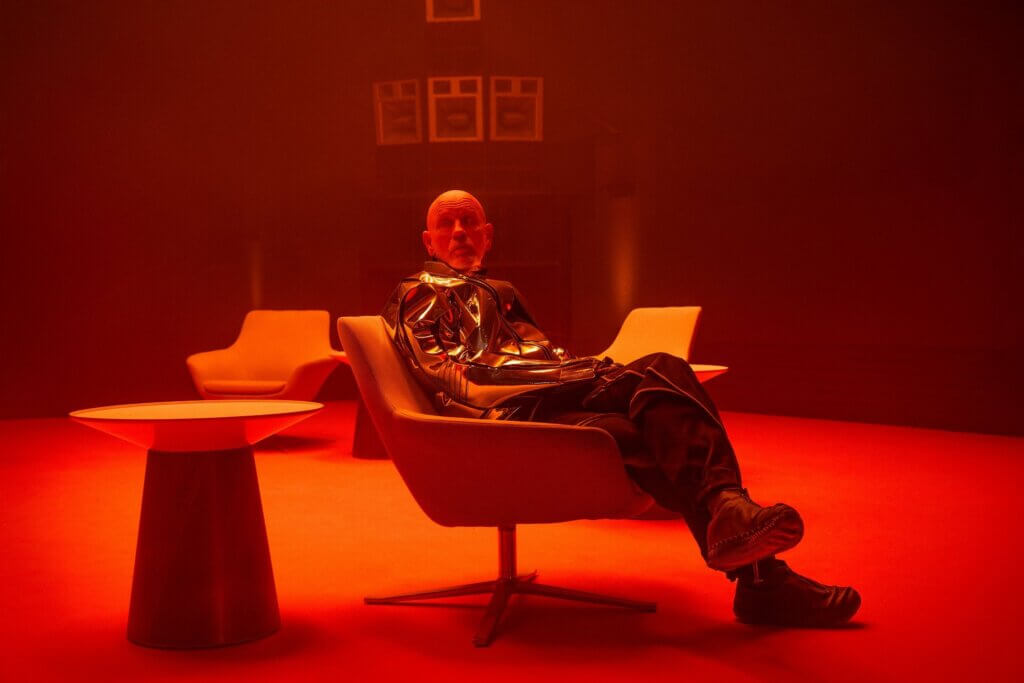Alfred Moretti sits beneath red light at his compound in Opus (2025)