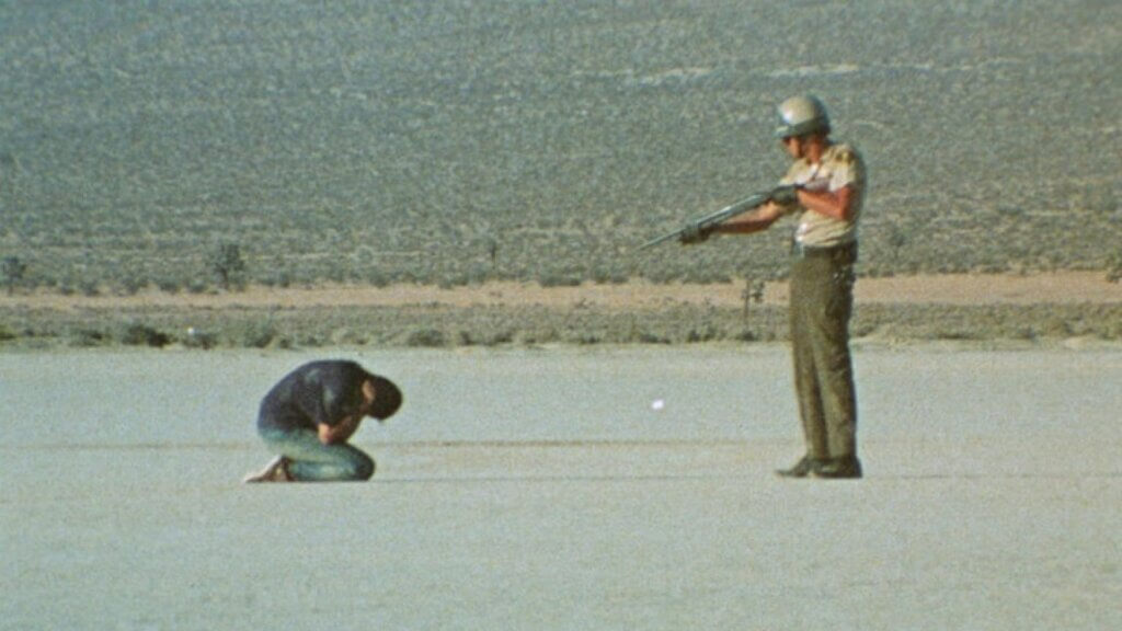 An armed officer points a rifle at a kneeling detainee in the desert in Punishment Park.