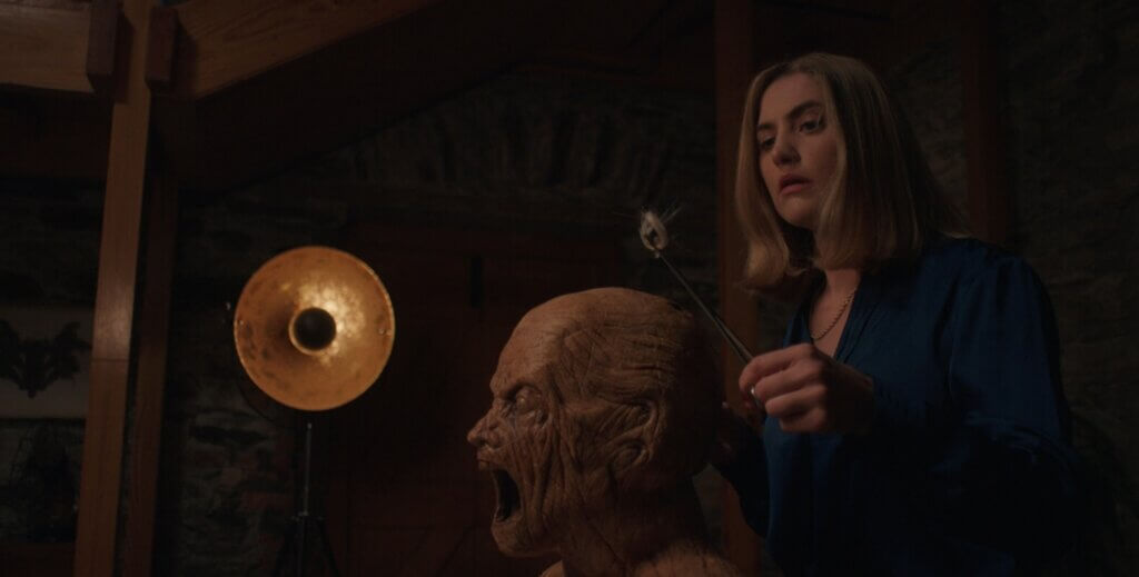 Yana holds a sharpened instrument beside the life-size wooden golem by lamplight in Oddity