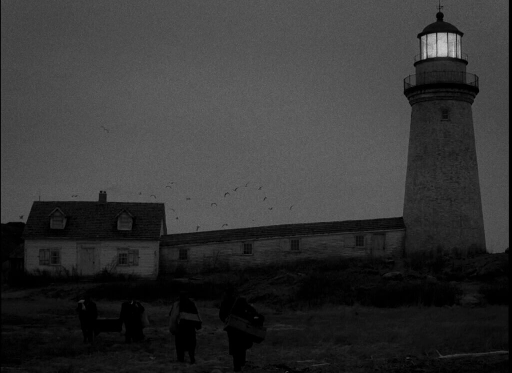 Two keepers carry supplies toward the island lighthouse in The Lighthouse (2019)