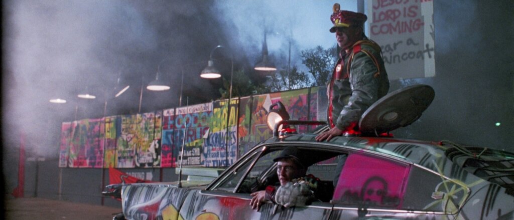 

A uniformed guard sits on a graffiti-covered car inside the neon-lit detention drive-in in Dead End Drive-In.