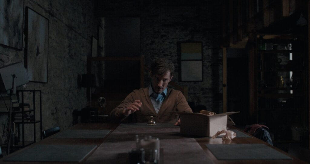 Ted sits alone at the long dining table inside the shadowy stone house in Oddity