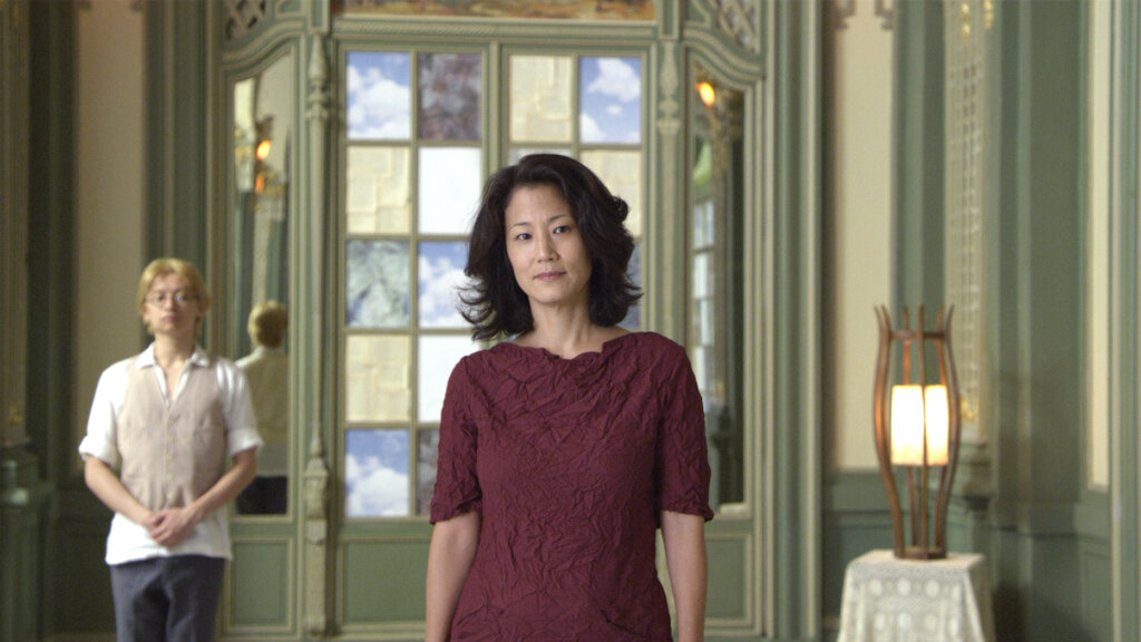 

Gwen stands in an ornate corporate hallway as a staff member waits behind her in Advantageous.