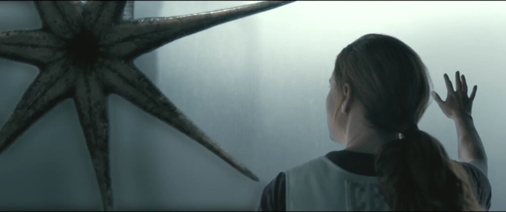 Louise Banks raises her hand toward a heptapod on the other side of the glass in Arrival.