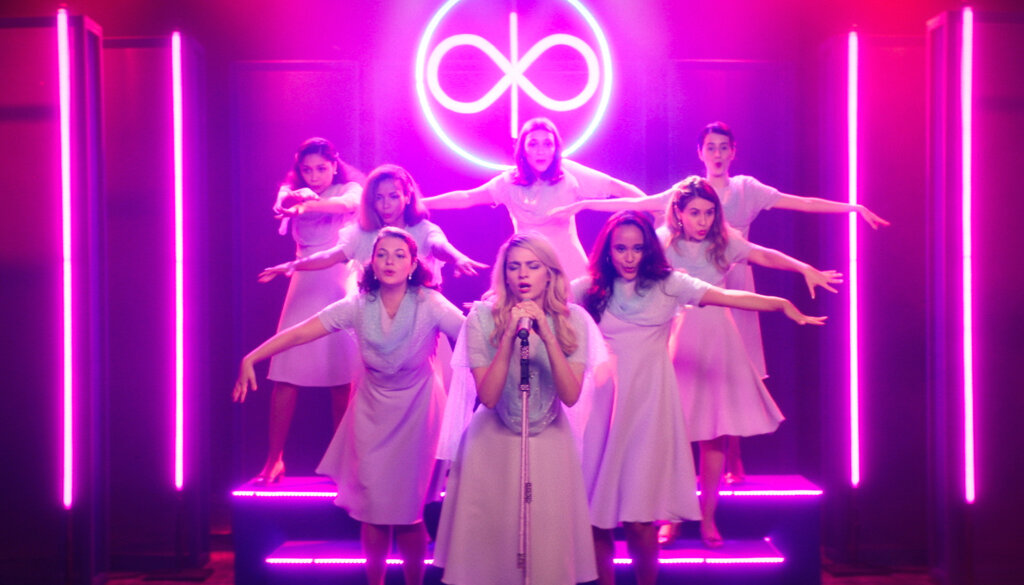 A female choir performs beneath magenta lights and a glowing symbol in Medusa.