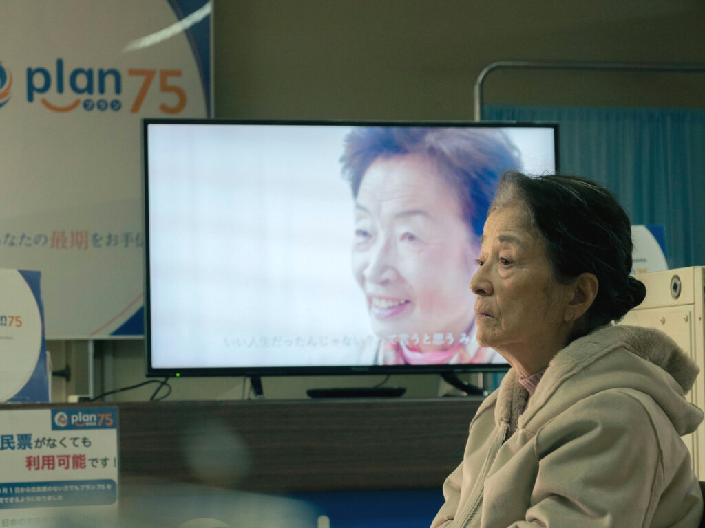 Michi sits beside a Plan 75 promotional display at a government service office in Plan 75.