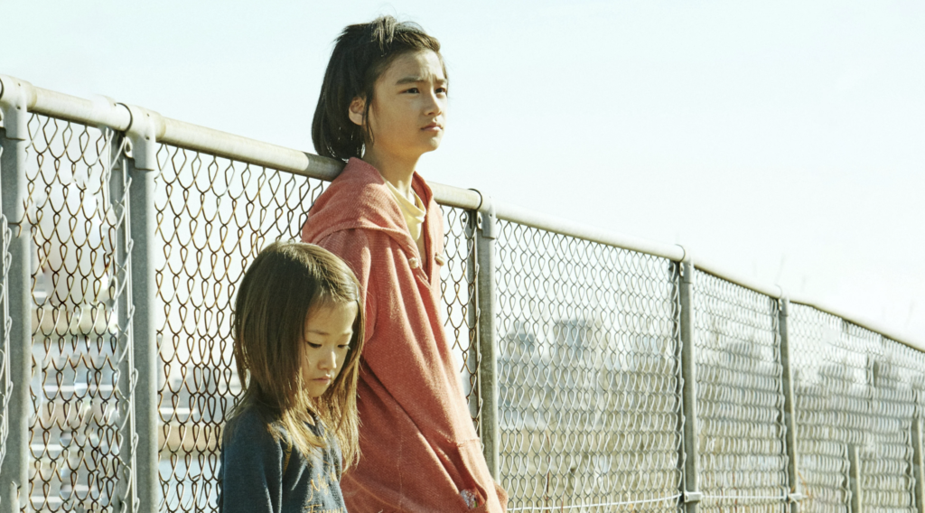 Shota and Yuri stand together beside a chain-link fence in Shoplifters (2018).