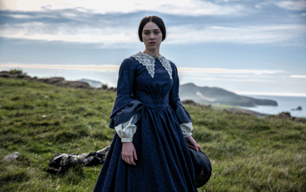 Ariāna Osborne stands on a grassy coastal hill wearing a blue Victorian dress in Mārama.