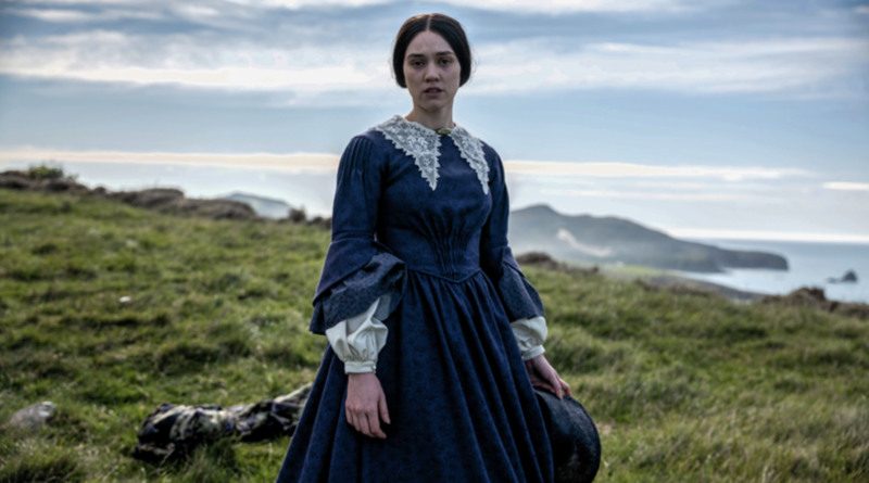 Ariāna Osborne stands on a grassy coastal hill wearing a blue Victorian dress in Mārama.