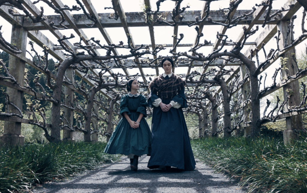 Mārama walks beside Anne beneath a vine-covered garden pergola in Mārama.