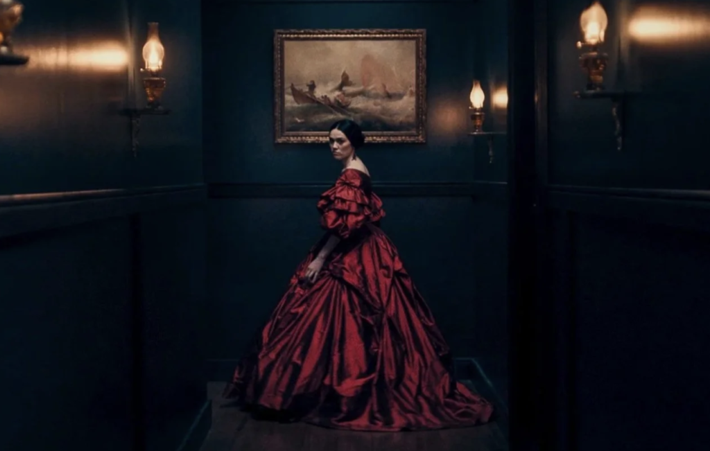 Mārama stands in the dark hallway of Hawkser Manor wearing an elaborate red Victorian gown.