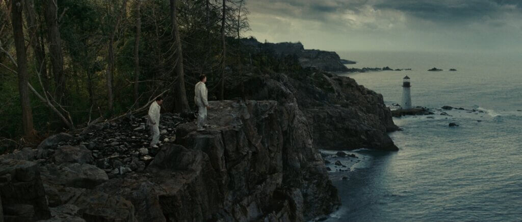 Two men in white stand on a rocky cliff above the lighthouse on Shutter Island.
