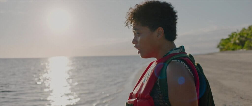 Jenn stands in profile on the beach wearing a red life vest as sunlight reflects across the ocean in Sweetheart (2019).