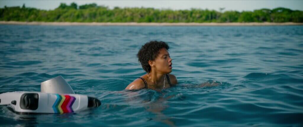 Jenn swims beside a floating suitcase with the island shoreline behind her in Sweetheart (2019).