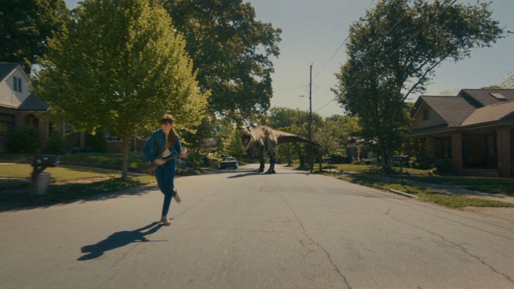 A dinosaur pursues a woman down the middle of a sunny suburban street in The End of Oak Street.