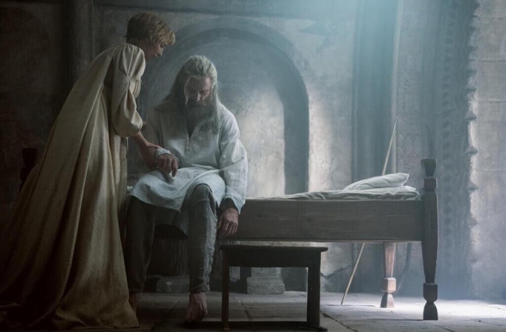 Brigid tends to a wounded Robin as he sits beside a bed in a stone chamber.