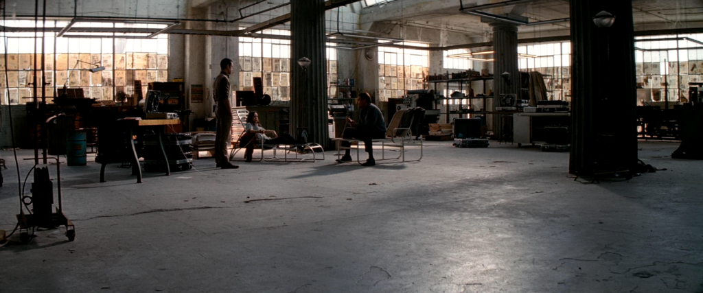 Arthur stands beside Ariadne and Cobb as they sit in reclining chairs inside the dream-sharing workshop in Inception (2010).