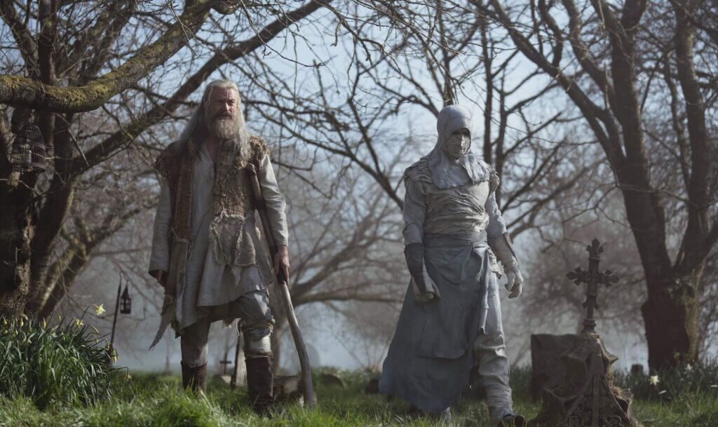 Robin walks through a graveyard beside a veiled leper.
