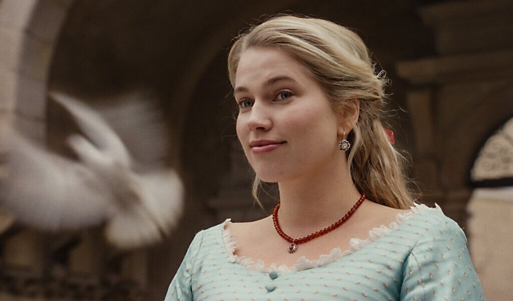 Agnes stands outdoors in a pale period dress.