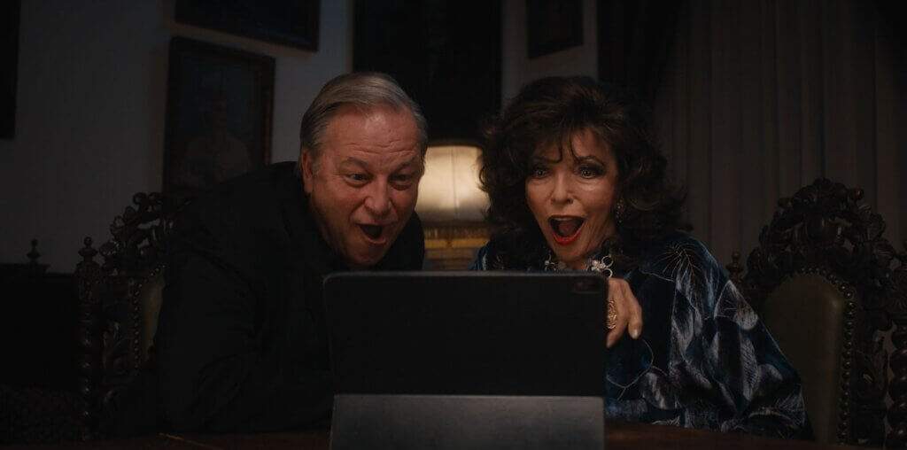 Francesca Carlyle (Joan Collins) and Chauncey (Jim Borstelmann) look at a laptop with surprised expressions in A Murder Between Friends (2026).