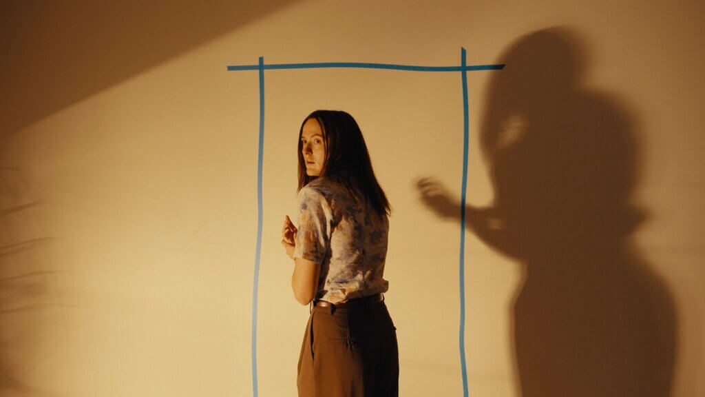 Mary stands within a blue tape doorway outline on a beige wall, with a large shadow beside her, in Backrooms (2026).