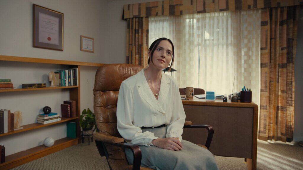 Mary sits in a brown leather chair beside bookshelves and a curtained window in Backrooms (2026).