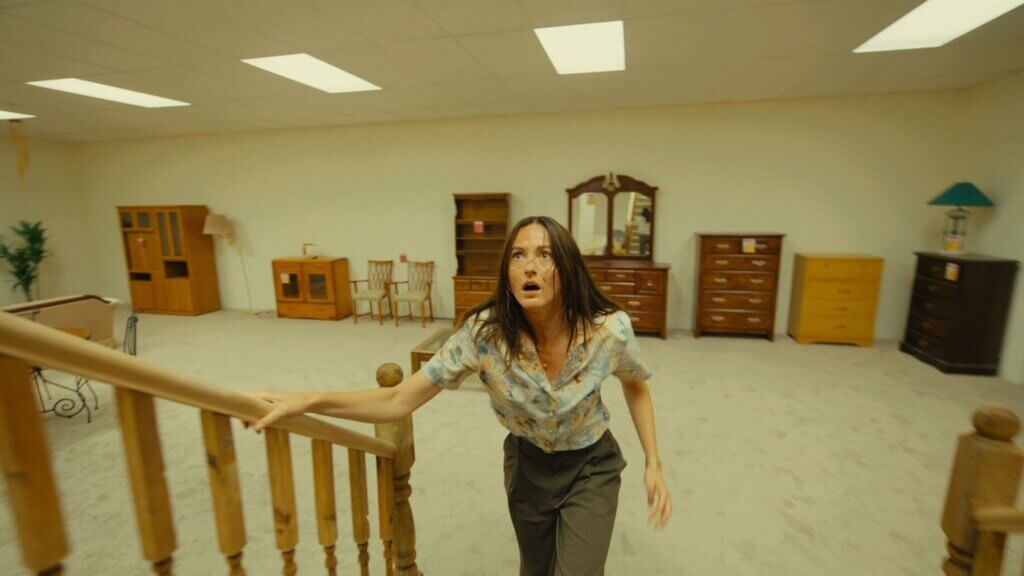 Mary grips a wooden stair railing in a yellow room filled with dressers and cabinets in Backrooms (2026).