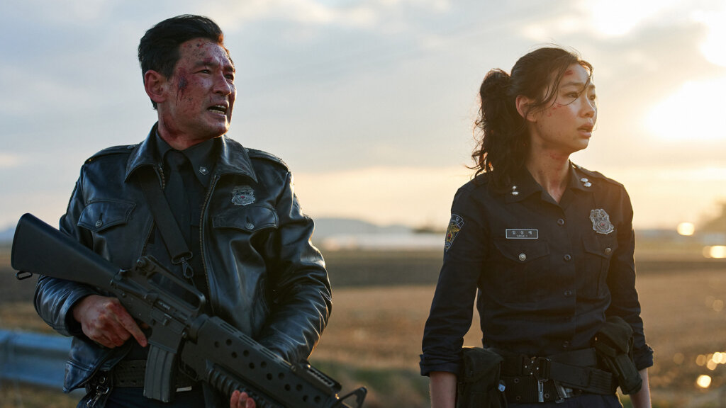 Bum-seok holds a rifle beside Sung-ae as the bloodied officers stand at sunset in Hope (2026).