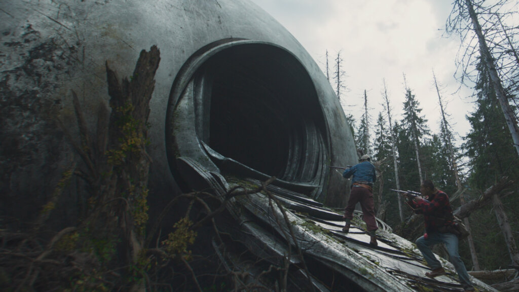 Two armed hunters approach the circular entrance of an alien spacecraft in a forest in Hope (2026).